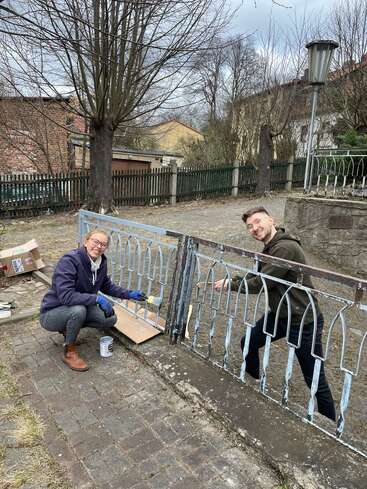 Two people are painting a metal fence outdoors. They are smiling, wearing casual clothes, and using brushes. The background shows trees, houses, and a cloudy sky.