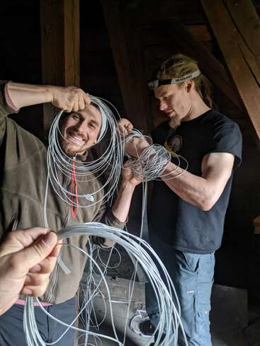 Two men stand indoors in a wooden room, smiling while untangling or handling coiled wires. One wears a headlamp, and a third hand holds more wire.