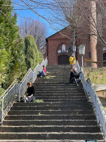 Four people sit separately on wide outdoor steps leading to a brick building. It’s sunny, trees are leafless, and the atmosphere appears peaceful and reflective.