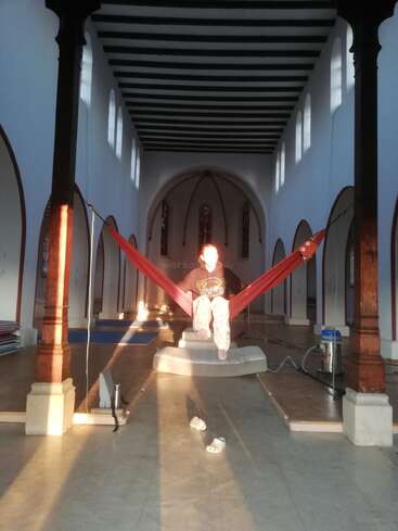 A person sits in a red hammock suspended between columns inside a large, sunlit church. Light shines on their face; slippers and a mattress below.