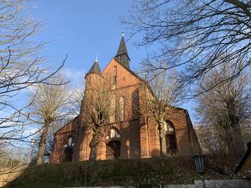 La imagen representa una majestuosa iglesia de ladrillo rojo con dos campanarios, enmarcada en un sereno cielo azul y rodeada de árboles sin hojas y exuberante vegetación.