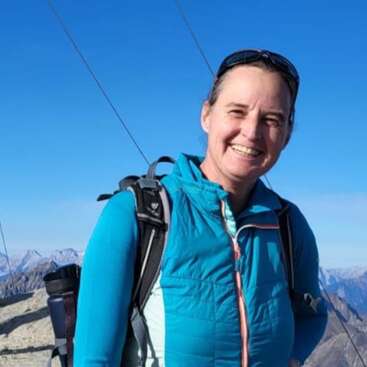 A woman smiles outdoors on a sunny day, wearing a blue jacket and backpack. Snow-capped mountains and a bright blue sky create a stunning background scene.