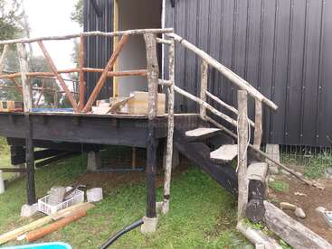 Das Bild zeigt eine rustikale Holztreppe mit Geländer, die zu einer kleinen erhöhten Veranda führt, die an ein schwarzes Gebäude angebaut ist, umgeben von Gras und Baumaterialien.