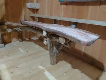 Auf dem Bild sieht man eine rustikale Holzbank, die ganz natürlich und unbearbeitet aussieht und in einem Raum mit Holzvertäfelung steht. Die Bank ist aus dicken, ungleichmäßigen Brettern gemacht.
