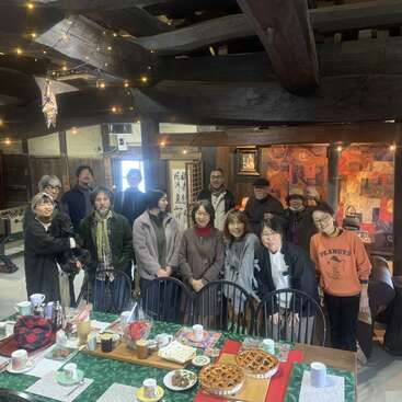 A group of people gathers indoors around a festive table with food. The cozy room features wooden beams, fairy lights, colorful paintings, and a foosball table.