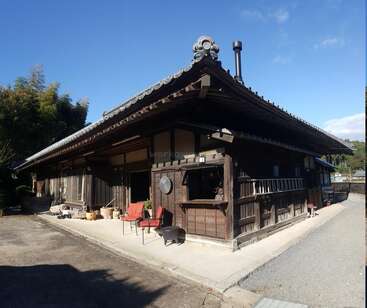 This image shows a traditional Japanese wooden house with a tiled roof, an open seating area, surrounded by trees, under a clear blue sky, on a sunny day.
