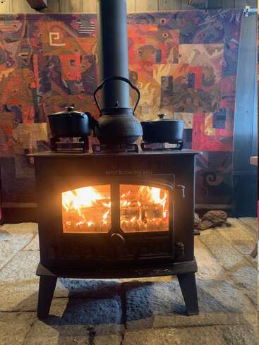 A cozy wood-burning stove with a visible fire warms the room. Three kettle pots sit on top; a colorful woven tapestry hangs behind, creating a homely atmosphere.
