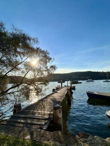 A wooden dock extends into a peaceful lake under a clear blue sky. Sunlight filters through tree branches, illuminating boats and hills in the tranquil distance.