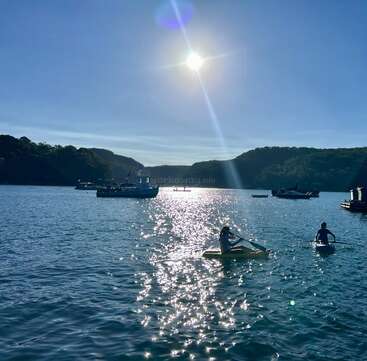 Under a bright sun, two people kayak on calm blue water, surrounded by boats and hills, with sunlight shimmering beautifully across the scenic bay.