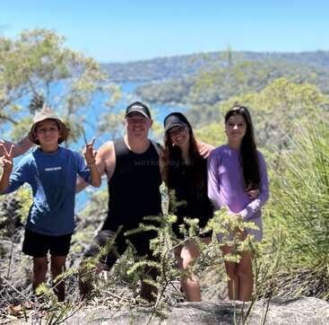 A family of four, smiling together in a sunny outdoor setting, stands surrounded by greenery with water and hills in the background, enjoying a nature hike.