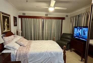 This cozy bedroom features a wooden-framed bed, pastel bedding, armchair, TV, artwork, ceiling fan, bedside lamps, large mirrored wardrobe, and heavy curtains.