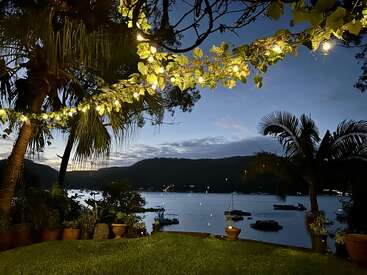 Fairy lights glow above a lush garden overlooking a calm lake at dusk. Palm trees frame the scene, boats rest peacefully on the water.