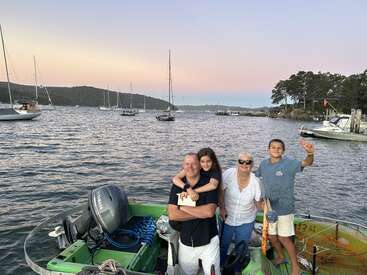A smiling family stands together on a boat at sunset, surrounded by calm waters and sailboats, with forested hills and a clear sky in the background.