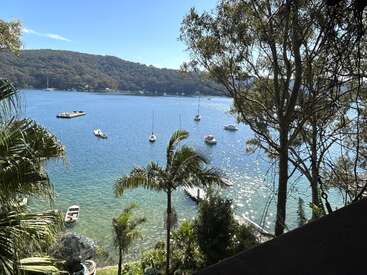 Sunlit blue water with small boats floating peacefully, surrounded by lush green trees and hills. Palm trees and dock in the foreground, creating a tranquil scene.