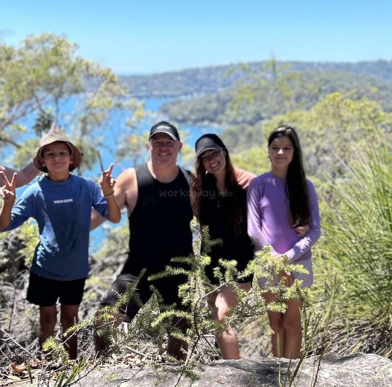A family of four, smiling together in a sunny outdoor setting, stands surrounded by greenery with water and hills in the background, enjoying a nature hike.