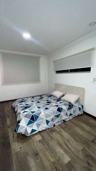 This image shows a simple, modern bedroom with a low bed, patterned bedspread, two pillows, white walls, wooden floor, and two large windows with blinds.