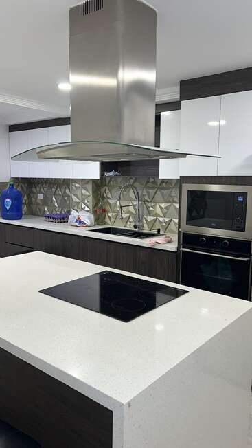 This modern kitchen features a sleek island with an electric cooktop, a stylish range hood, geometric backsplash, built-in oven, sink, and contemporary cabinetry.