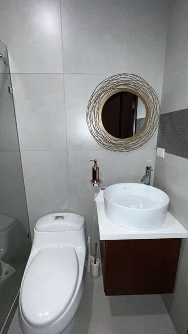 This modern bathroom features a sleek toilet, round vessel sink, stylish mirror with gold accents, dispenser, dark vanity, neutral tones, and minimalist décor, creating a clean, elegant space.