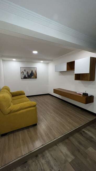 A modern, minimalistic living room featuring a mustard yellow sofa, abstract mountain wall art, floating wooden shelves, and laminate wood flooring with white walls.