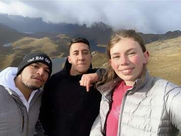 Three friends pose for a selfie in a mountainous landscape, smiling and bundled up in jackets, with misty peaks and grassy hills in the background.