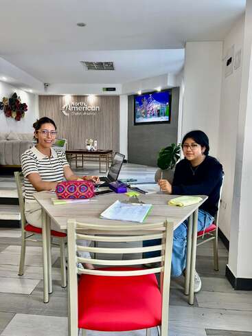 Two women are sitting at a table with laptops and notebooks, studying or working together at North American English Academy. A TV and decor are visible.