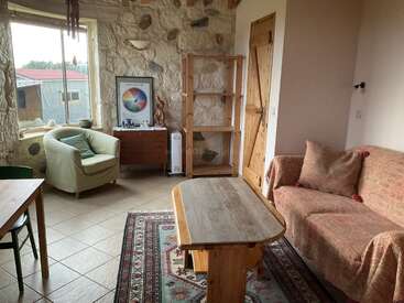 This cozy room features a green armchair, wooden table, patterned rug, rustic stone walls, empty bookshelf, framed art, and a beige couch with cushions.