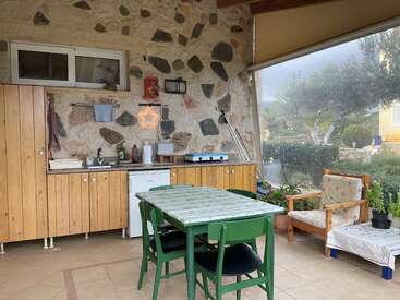 This cozy outdoor kitchen features wooden cabinets, a small fridge, a stove, green dining table, cosy seating, stone walls, and looks out onto a garden.
