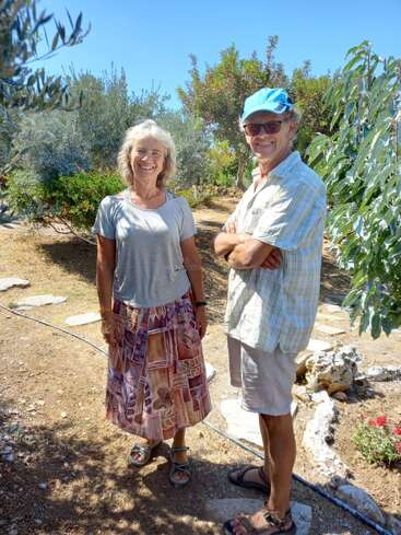 A smiling woman and man stand together in a sunlit garden, surrounded by trees and plants, wearing casual summer clothes and sandals, enjoying a beautiful day.