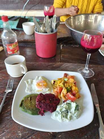 Ein rustikaler Tisch, gedeckt fürs Frühstück, mit einem bunten Teller voller Essen, Kaffee, Besteck, einer großen Salatschüssel, Weingläsern mit einem roten Getränk und Soße.