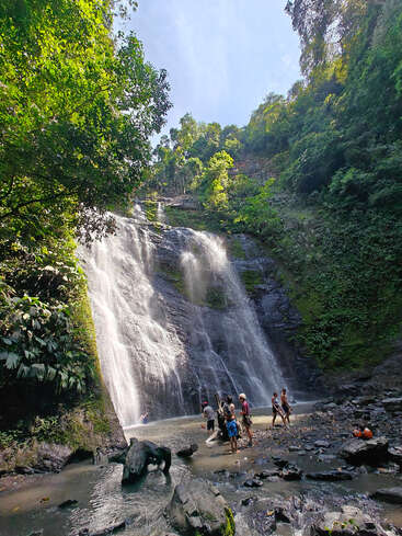 Eine Gruppe Leute steht am Fuße eines atemberaubenden Wasserfalls, umgeben von üppigem, grünem Wald. Das Sonnenlicht fällt durch die Bäume und schafft eine faszinierende, natürliche Kulisse.