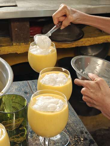 Jemand löffelt eine weiße, cremige Garnierung auf Gläser, die mit einem dicken gelben Smoothie oder Dessert, vielleicht Mango, gefüllt sind, auf einem rustikalen Holztisch.