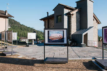Múltiples fotografías artísticas expuestas al aire libre en soportes, cerca de una rústica casa de piedra en un pintoresco pueblo de montaña, crean un ambiente de galería al aire libre.