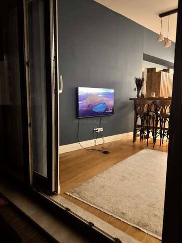 A modern living space with wooden floors features a mounted TV, a rug, bar stools, and pendant lights, viewed from an open sliding glass door. Relaxing atmosphere.