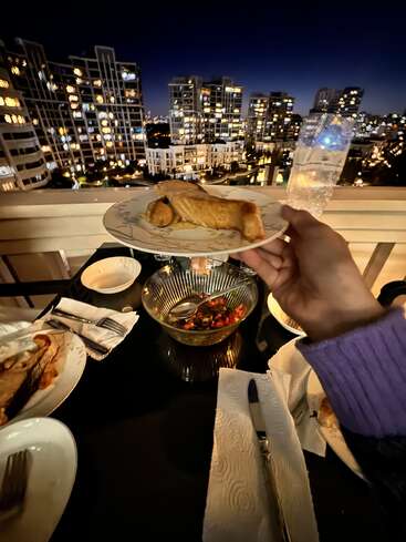 Uma pessoa segura um prato com comida em uma varanda à noite, com vista para as luzes da cidade e prédios altos, desfrutando de um jantar ao ar livre com amigos.