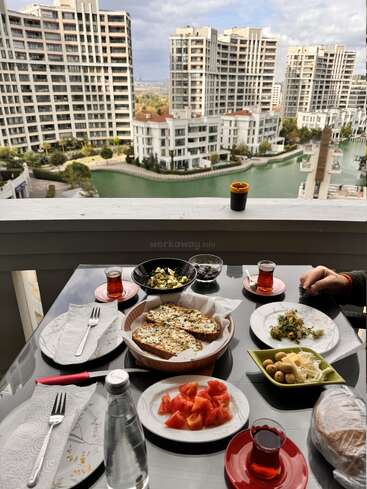 Uma mesa de café da manhã com chá, pão, queijo, azeitonas, tomates e salada tem vista para um canal panorâmico cercado por modernos prédios de apartamentos.
