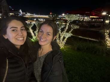 Dos mujeres que sonríen mientras se hacen un selfi por la noche, rodeadas de árboles iluminados y luces navideñas, con un pintoresco paseo marítimo y un restaurante al aire libre al fondo.