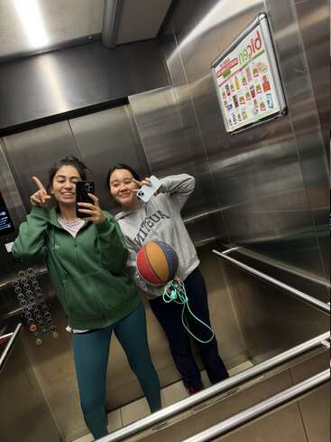 Dos chicas se hacen una alegre selfie frente al espejo dentro de un ascensor. Una sostiene un teléfono, la otra lleva una pelota de baloncesto y una cuerda para saltar. Ambas tienen un aspecto deportivo y están sonriendo.