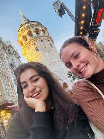 Dos jóvenes sonríen para hacerse un selfi frente a la histórica Torre de Gálata en Estambul, rodeadas por los edificios de la ciudad, las luces y un cielo azul brillante.