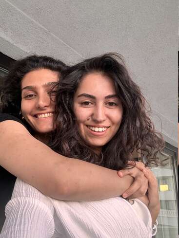 Dos jóvenes sonríen con calidez mientras se abrazan con fuerza. Parecen felices y relajadas, de pie juntas al aire libre, con luz natural y un muro de hormigón a sus espaldas.