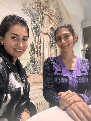 Dos mujeres sonríen sentadas a una mesa en el interior, con un mural artístico muy detallado que representa arquitectura clásica y árboles al fondo. Ambiente cálido y acogedor.