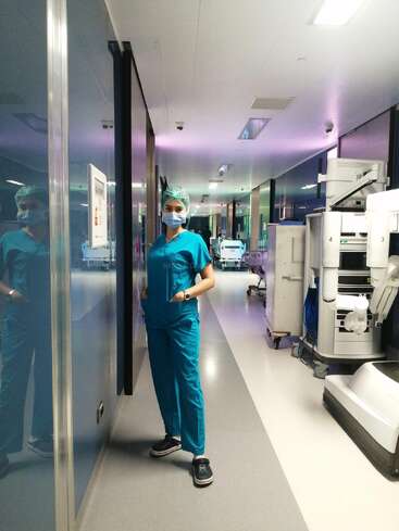 Un profesional sanitario, vestido con una bata azul y una mascarilla, se encuentra en el pasillo de un hospital repleto de material médico, irradiando una tranquila seguridad en sí mismo y disposición para el servicio.