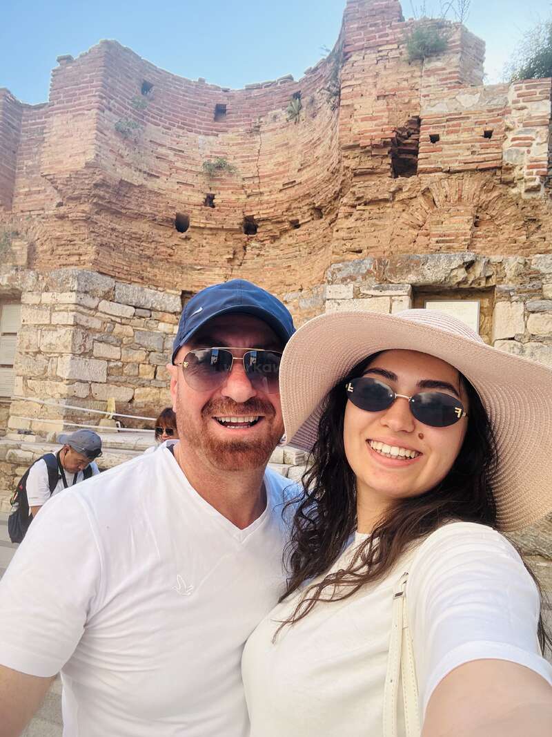 Um casal sorridente, usando óculos escuros e chapéus, posa para uma selfie em frente a antigas ruínas de tijolos, aproveitando um dia ensolarado de turismo com outros turistas nas proximidades.