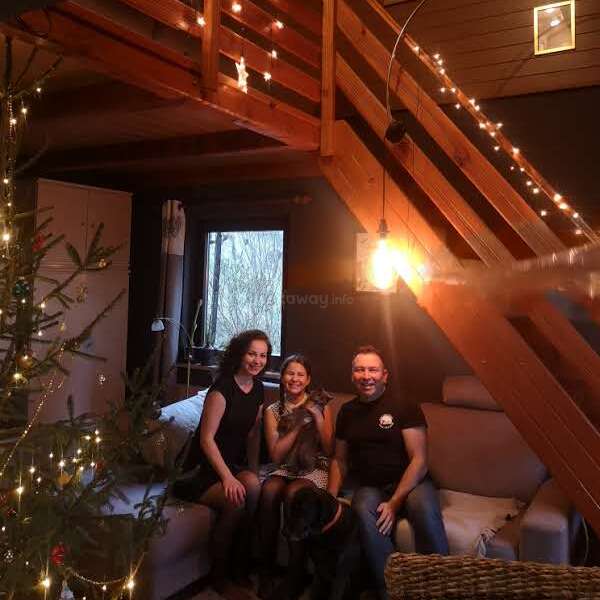 Tres personas y un perro están sentados alegremente en un sofá, en una habitación acogedora e iluminada con una cálida luz, decorada con adornos festivos, como guirnaldas de luces y un árbol de Navidad.
