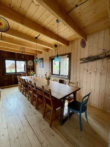 A cozy wooden dining room with a long table, mismatched chairs, pendant lights, and flower vases. Windows and handcrafted decorations add warmth and rustic charm.
