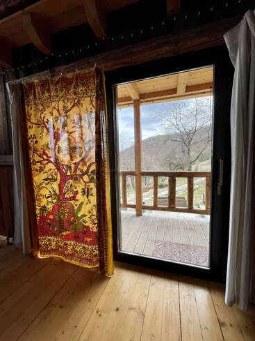 The image shows a cozy wooden room with a decorative yellow curtain, a glass door opening to a porch, and scenic mountain and tree views outside.