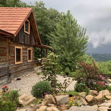 A rustic wooden cabin with a red roof stands beside a colorful garden. Trees surround the house, and dark storm clouds loom over distant green hills.
