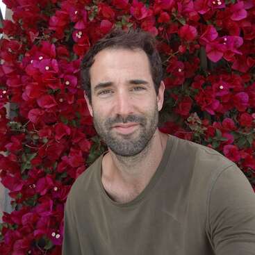 Un homme aux cheveux bruns et à la courte barbe sourit, posant devant un mur vibrant rempli de fleurs rouges luxuriantes, vêtu d'une chemise verte.
