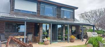 Esta imagem mostra uma casa rústica de dois andares com revestimento de madeira, janelas grandes, um pátio coberto, móveis externos, vasos de plantas e um quintal bem cuidado.