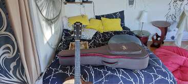 Um quarto aconchegante com uma colcha estampada em preto e branco, travesseiros amarelos, violão em um estojo, mesas laterais circulares, arte na parede e janelas com cortinas.