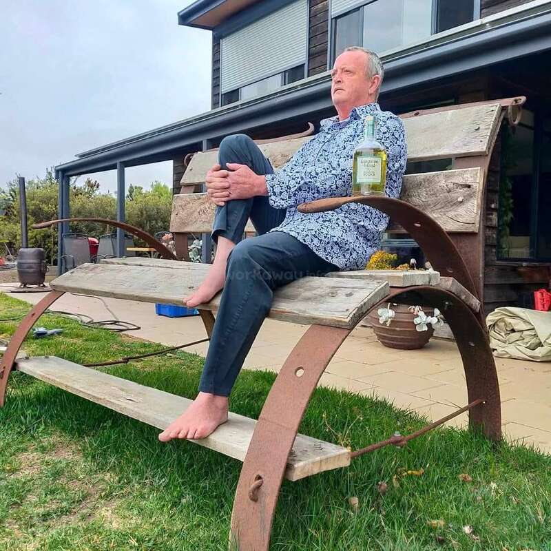 Um homem senta-se descalço em um banco de madeira de grandes dimensões em um jardim, segurando uma garrafa e parecendo pensativo. Uma casa moderna e vegetação estão atrás dele.